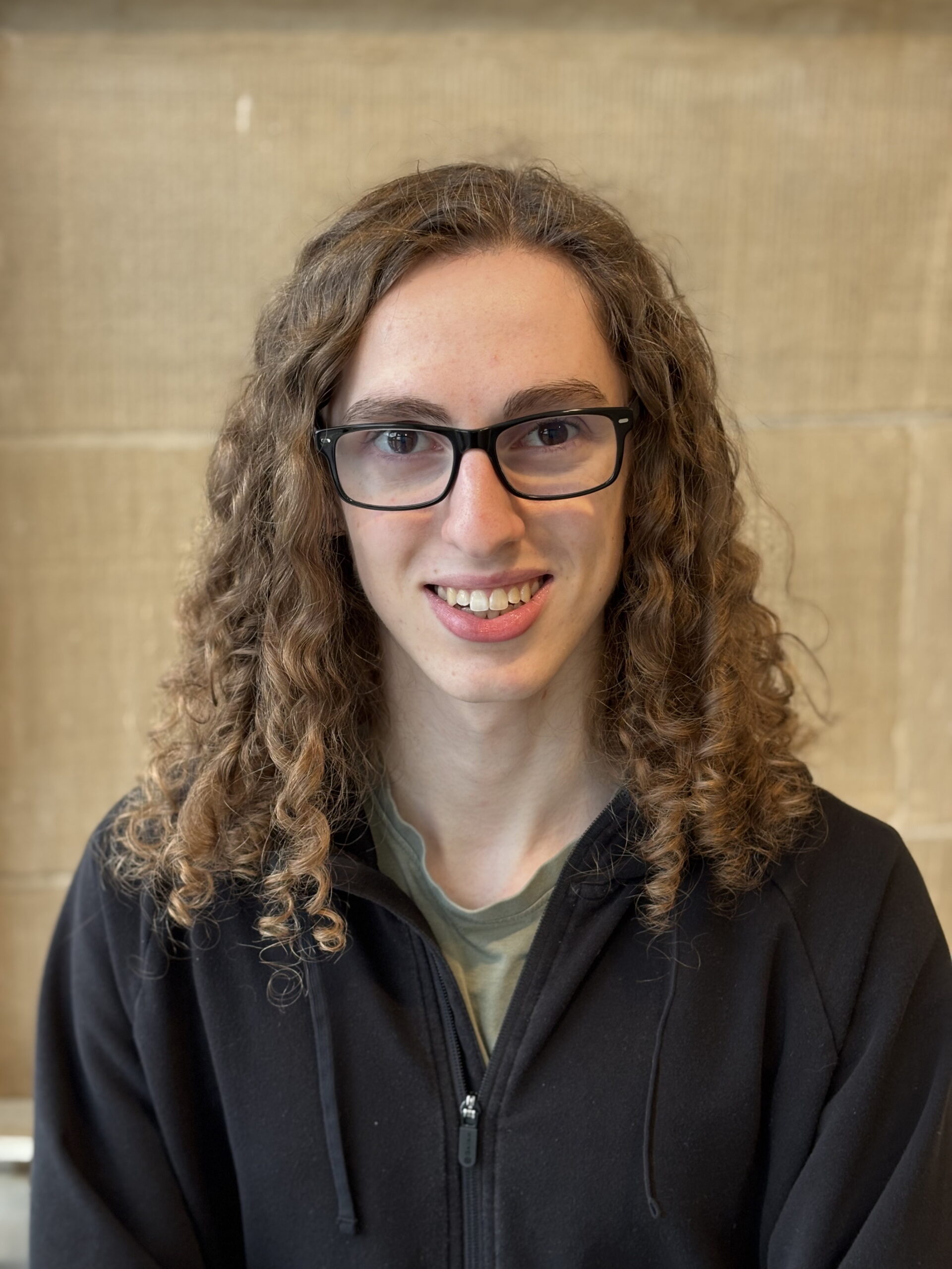 Headshot of Connor Bouwens. Connor has long, light brown curly hair and wears dark framed glasses. He's wearing a sage green t-shirt under a black partially unzipped hoodie.